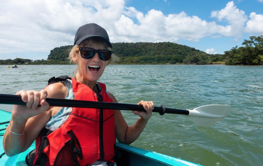 An older adult wearing polarized sunglasses smiles and holds up their paddle as they kayak across blue waters.