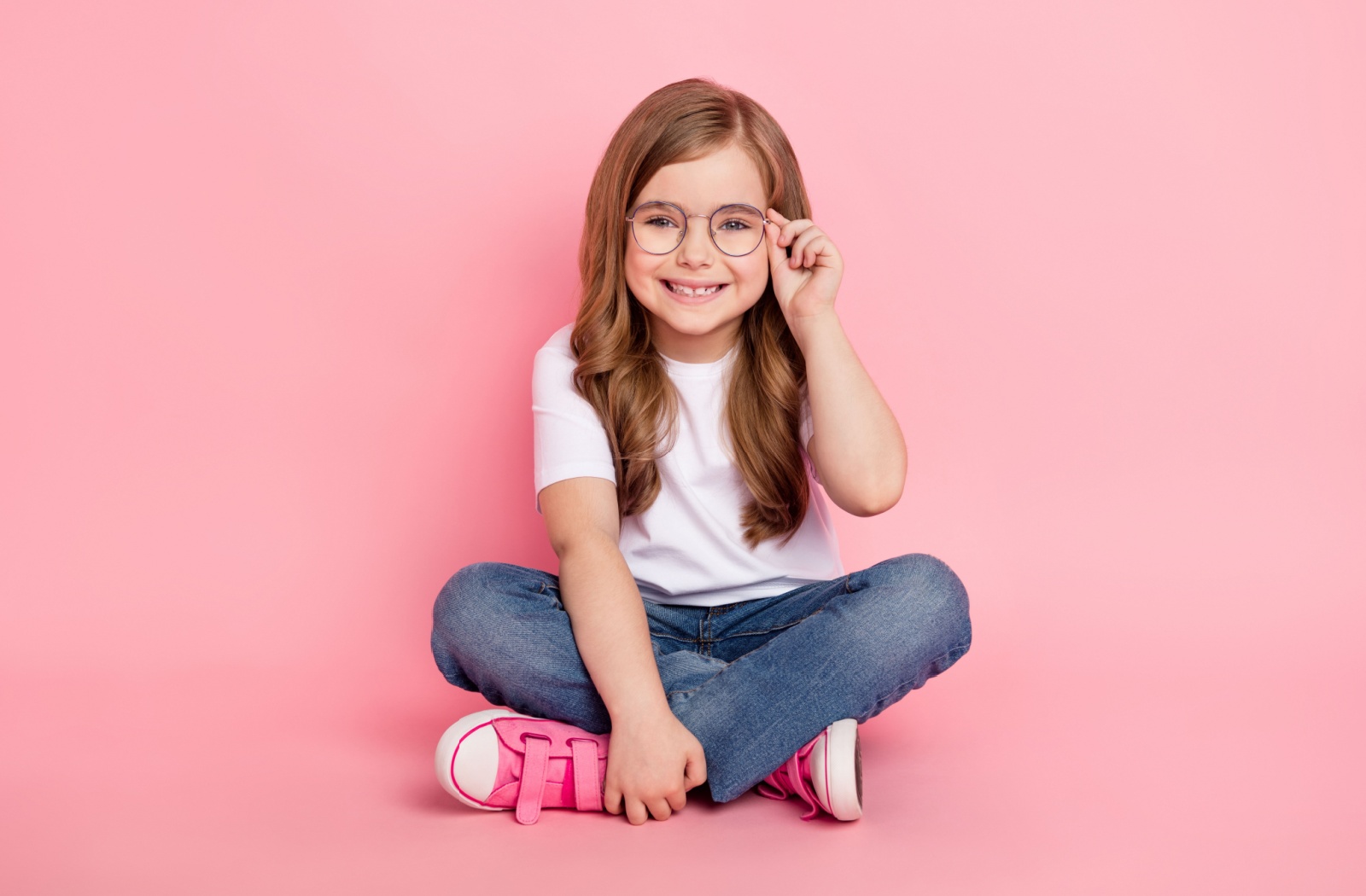 Young happy girl sitting cross legged against pink background wearing circular glasses.