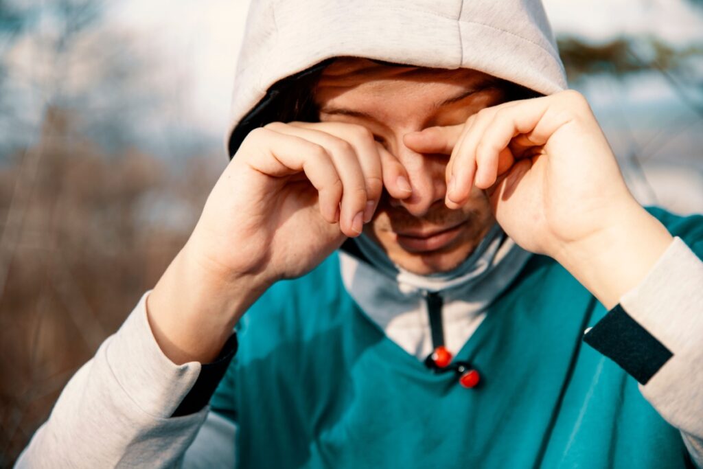 Person wearing winter hoodie outdoors rubbing irritated eyes, showing dry eye discomfort caused by cold air, low humidity, and harsh winter weather conditions affecting tear film.