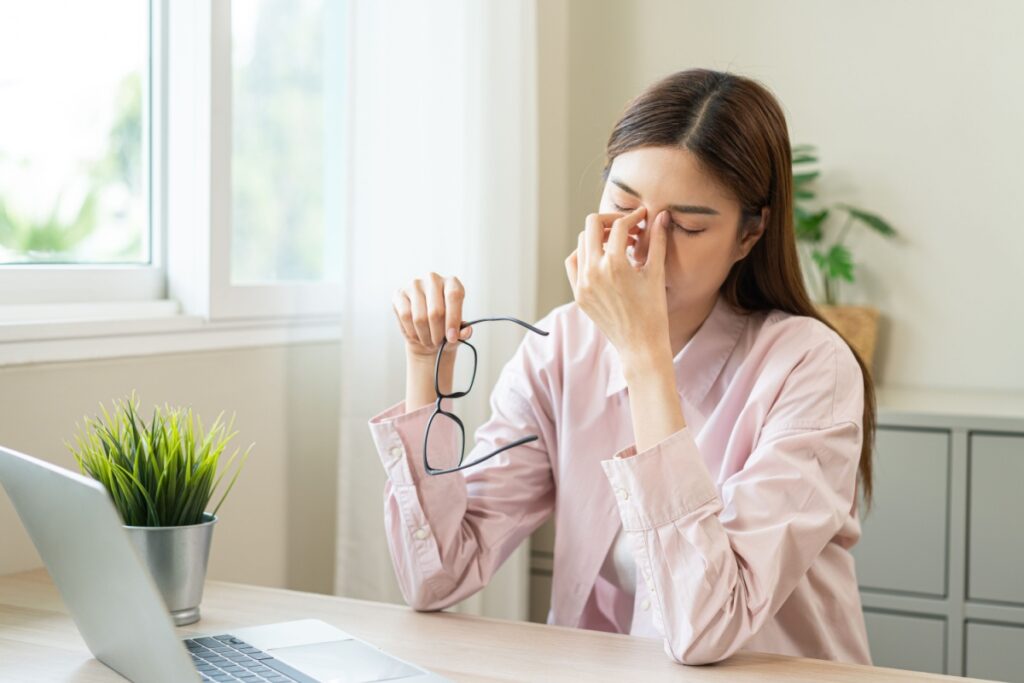 Woman at desk removing glasses and rubbing tired, dry eyes while working on laptop, demonstrating digital eye strain and dry eye symptoms from prolonged screen time and indoor heating during winter months.