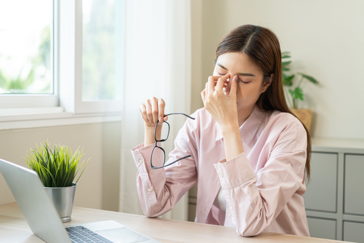Woman at desk removing glasses and rubbing tired, dry eyes while working on laptop, demonstrating digital eye strain and dry eye symptoms from prolonged screen time and indoor heating during winter months.